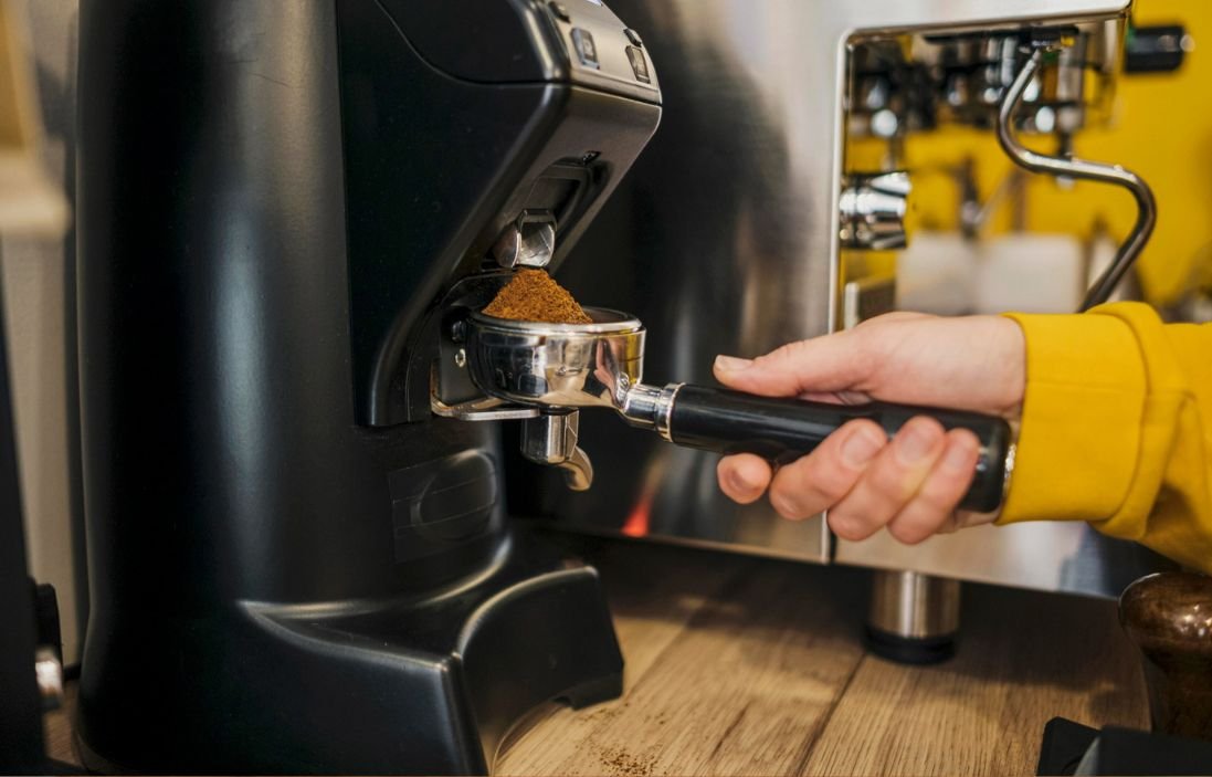 A person in a yellow sleeve holds a portafilter under a grinder dispensing coffee grounds. A coffee machine and wooden counter are visible.